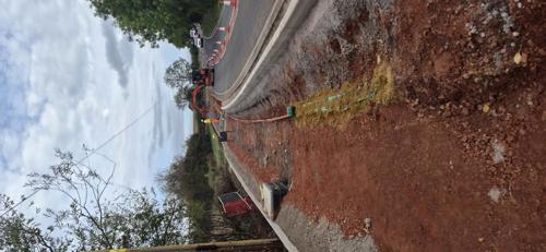 Construction of a new shared-use path alongside a curved roadway. The image shows a newly laid kerb separating the path from the road, with exposed red earth and excavation works in the foreground. An excavator and road workers in high-visibility clothing are visible in the distance, while traffic passes through a coned-off lane management system. Trees and vegetation line both sides of the route under an overcast sky.