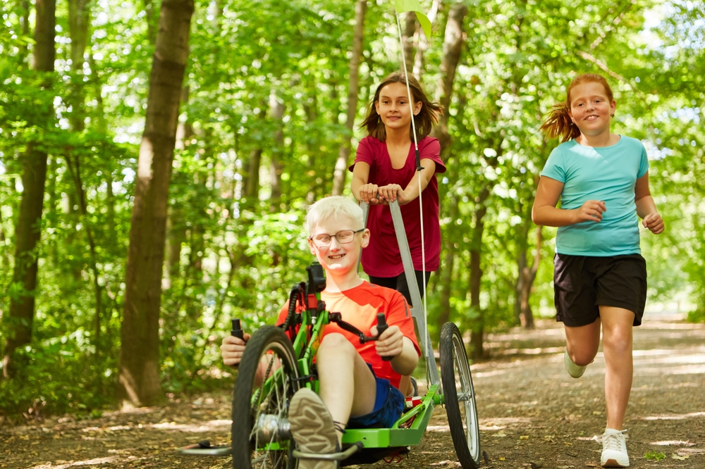 Three children in a forest in the high summer with one using a recumbent cycle
