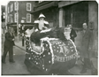A black and white photo of a Christmas sled on the roads of a town, with some formally dressed men walking around it.