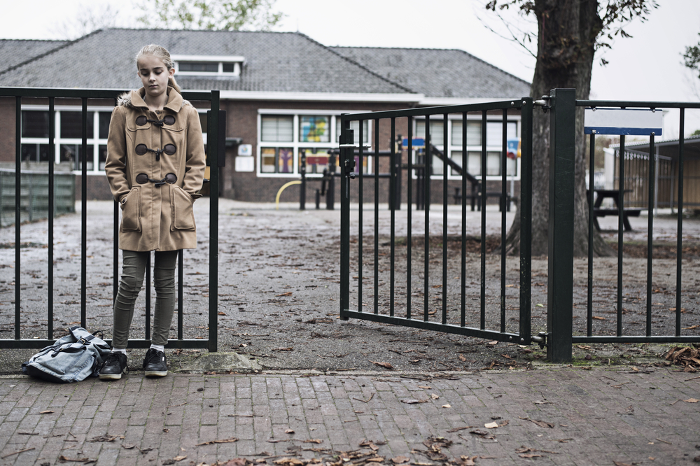 Sad girl with her hands in her pockets standing in front of school gate