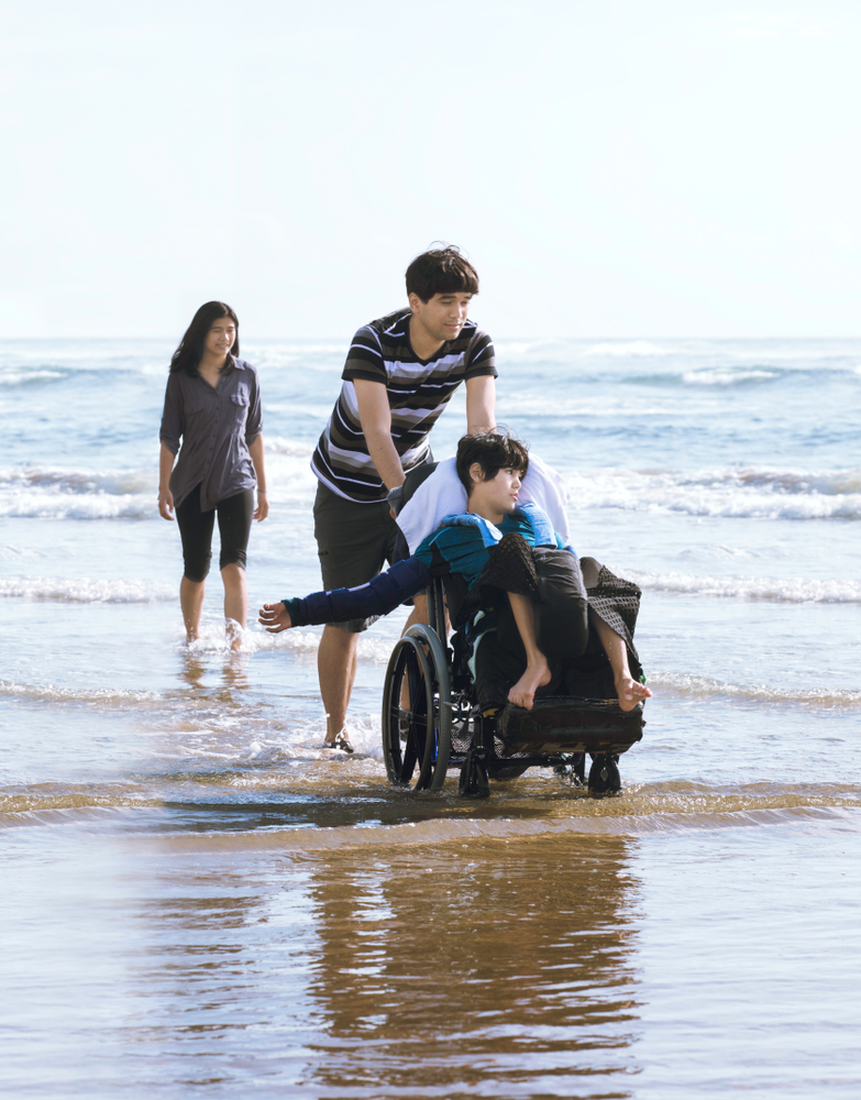 Three people by the water’s edge at a beach moving along, two on foot and one on a wheelchair