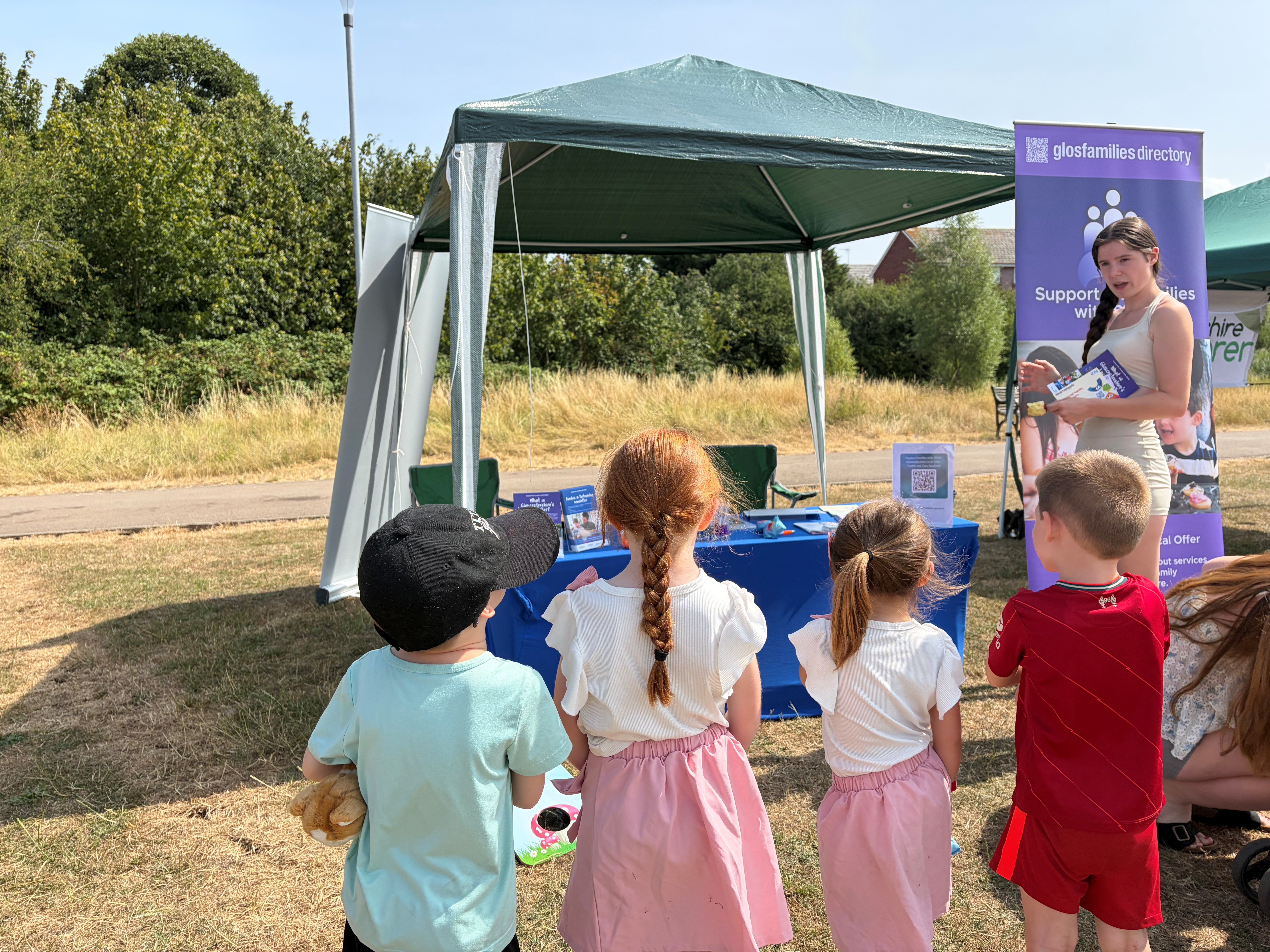 Four children and a Parent Carer visiting the SEND Local Offer stand at a family fun day - children queuing to play bean bag toss and Parent Carer is holding some leaflets she has picked up from the stand