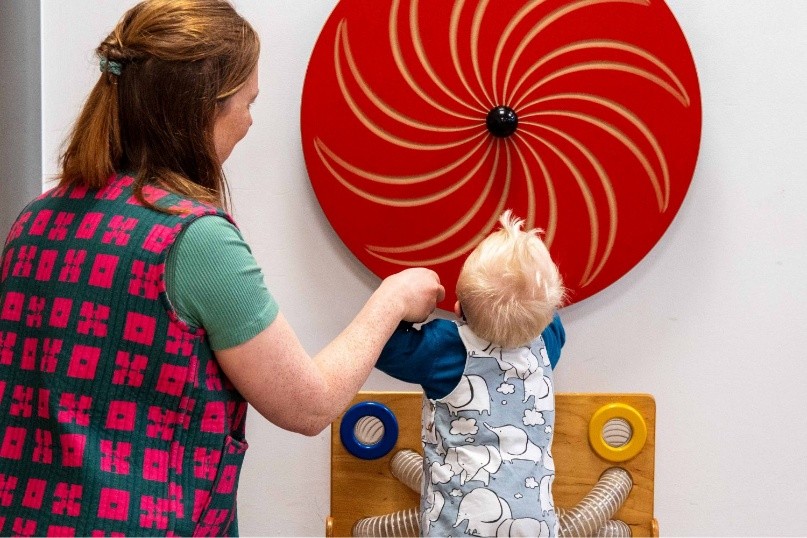 A parent supports a young child during a play-based activity, exploring a large red sensory spinner mounted on the wall as part of an early years session.