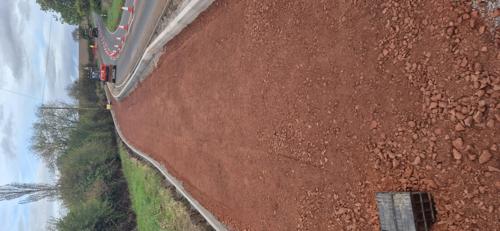 A newly constructed section of shared-use path stretches alongside a rural road, with a compacted red stone surface and new kerbs installed on both sides. An excavator is positioned further along the route, while traffic passes through a coned-off lane under temporary traffic management. Grass verges, hedgerows and trees line the path, and the route follows a gentle curve through the countryside beneath a cloudy sky.