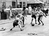 A black and white photo of some children playing football in the middle of the road.