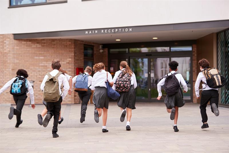 A mixed group of eight students with their backs to the camera wearing a grey school uniform running towards entrance doors to a school building with the words ‘Main reception’ above the doors