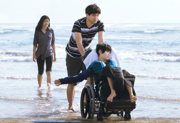 Three people by the water’s edge at a beach moving along, two on foot and one on a wheelchair