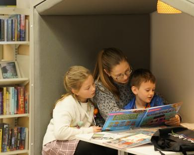 Parent carer with a boy aged four and a girl aged seven reading a book together  in an enclosed seating area in Stroud library