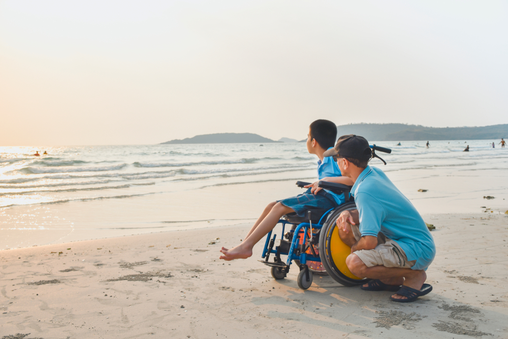 Young boy in a wheelchair on a beach looking out to sea with his carer crouching down beside the chair also looking out to sea