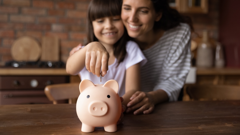 Mother hugs preteen daughter who is dropping coins into piggybank to save money. 