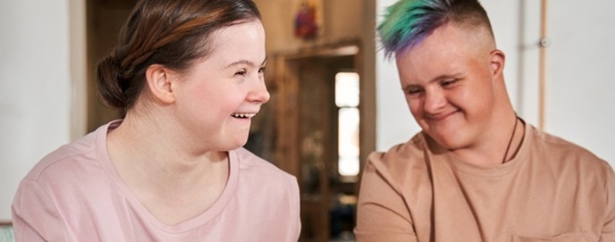 Two young adults sit adjacent in a recreational setting. Both are wearing casual clothing and are smiling