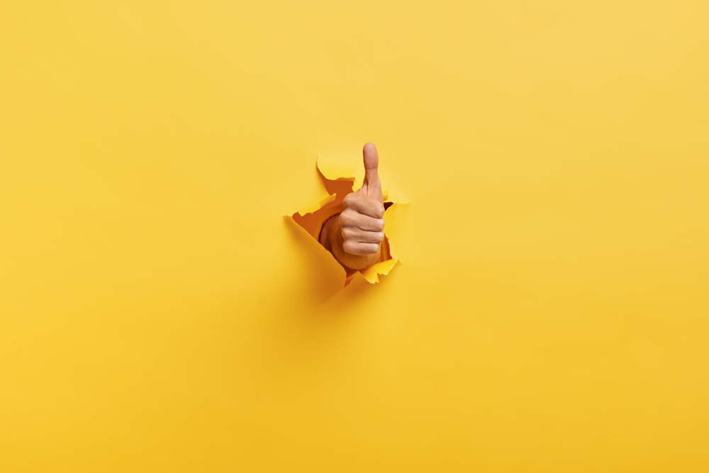 Yellow background with a fist punched through it showing a thumbs up approval sign