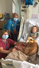 A child in a hospital bed with two nurses next to the bed and a dog on the girls lap
