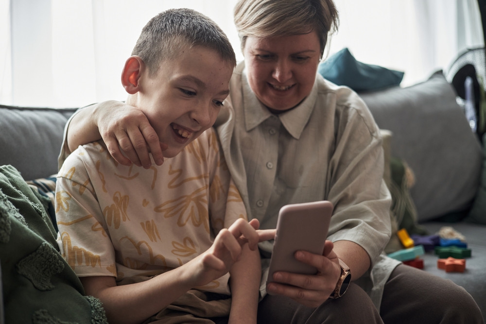 Portrait of a young man smiling with an adult and being supported to use a smartphone.