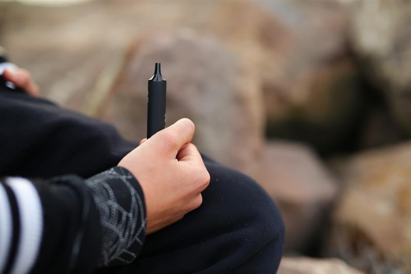 A young person  of thirteen years old, holds a vape in his hands. A teenager smokes a disposable electronic cigarette while sitting  