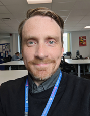A photo of Dan Furmage. He has short light brown hair and blue eyes, and is smiling at the camera. He is stood in a classroom.