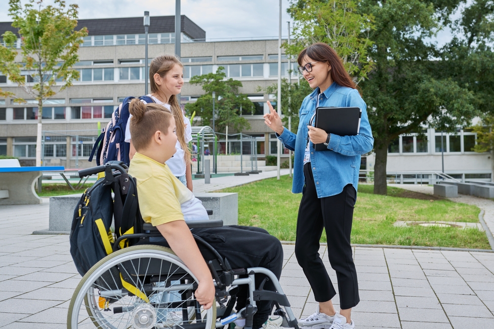 Two older pupils, one in a wheelchair outside a large school being greeted by a teacher