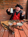 A photo of a man wearing a pirate hat and sat at a table with empty food plates surrounding him. He is grinning and holding a tankard.