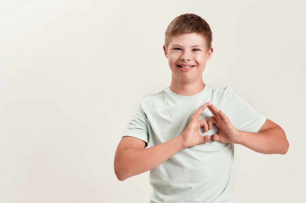 Smiling young boy in a pale green t-shirt making a heart symbol with his hands against his chest.