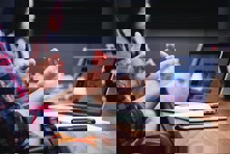 A person wearing a red and blue plaid shirt is gesturing with their hands while sitting at a wooden table. In front of them is an open laptop displaying colorful icons or images, alongside an open notebook with a smartphone on top. Another person is partially visible in the blurred background, suggesting a meeting or discussion in an office setting