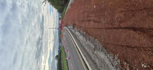 A newly constructed shared-use path under development runs alongside a rural road. The path surface is compacted red stone, bordered by new kerbs separating it from the carriageway. Traffic management is in place with cones lining the road and temporary barriers visible ahead. Grass verges and hedgerows flank the route, while a cloudy sky stretches overhead. The image shows the route taking shape before the final surface is laid.