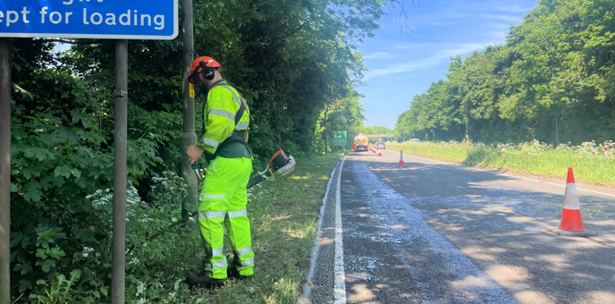 Photo of person wearing high visibility protective clothing and using a strimmer to cut a grass verge on a sunny day.