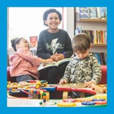 Three children are seated in a library setting with shelves of books in the background. One child is holding an open book, while another smiles at him and is touching the book. The other child is engaged with colourful bead maze toys on a table in front of them. 