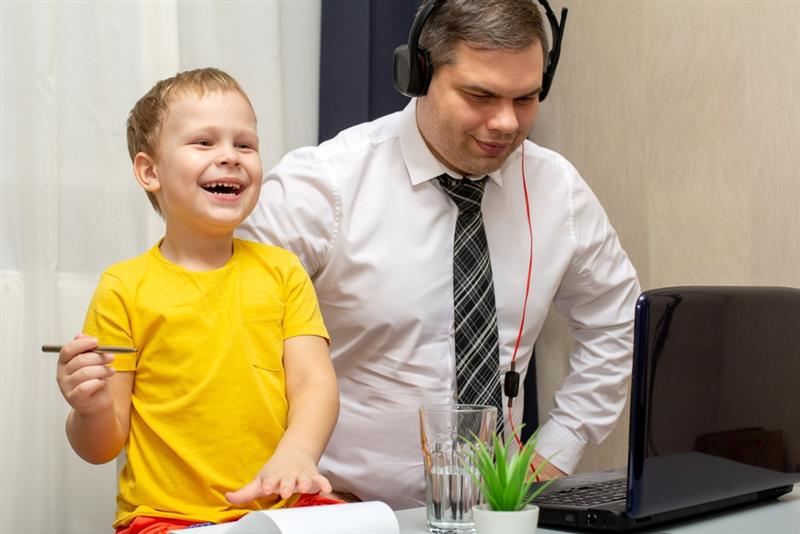 Boy aged four is smiling and wearing a yellow t-shirt and holding a pencil sharing a desk with a man wearing a shirt, tie and a headset whilst working on a laptop. 