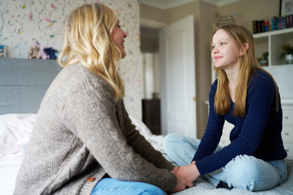 Woman sitting on a bed with an older teenage woman holding hands and smiling