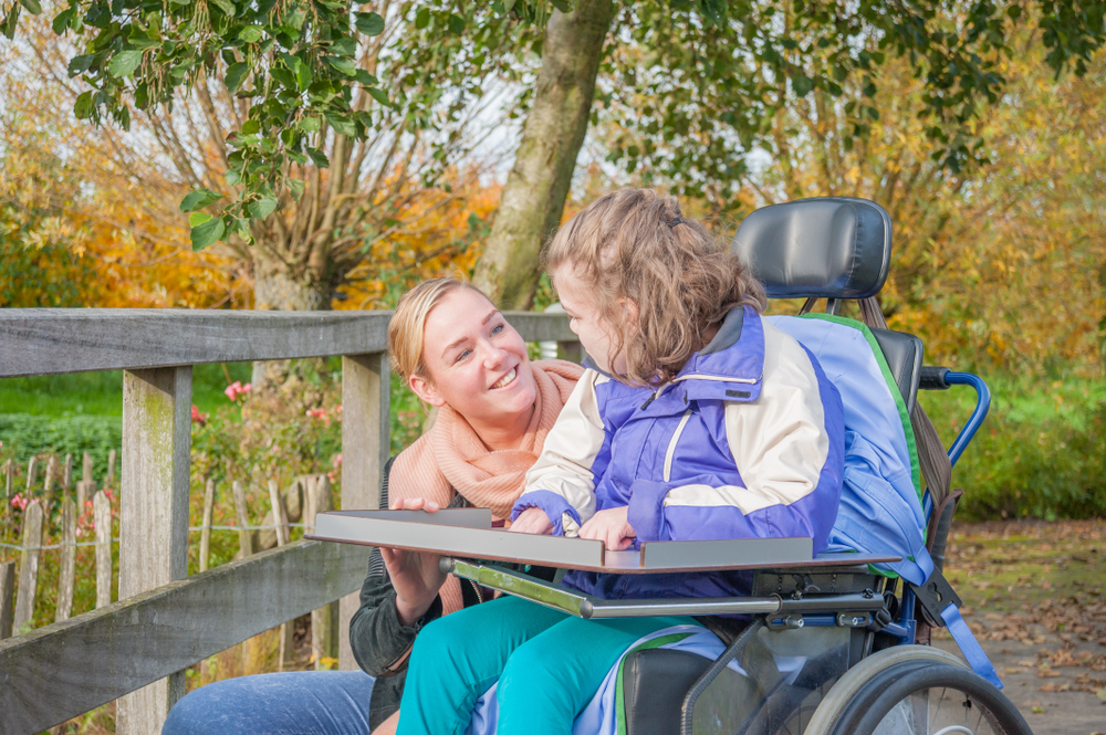 A Carer Talks To A Person She Is Caring For Who Is In A Wheelchair Next To A Field 