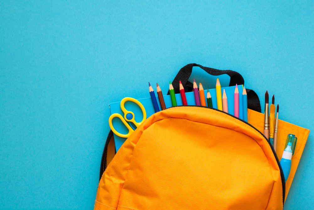 A yellow rucksack on a jade background with the zip open showing a selection of stationery items